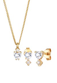 Ensemble de bijoux dorés comprenant un collier à chaîne avec un pendentif en forme de cœur et des boucles d'oreilles à clou assorties en forme de cœur, ornés de pierres précieuses transparentes.