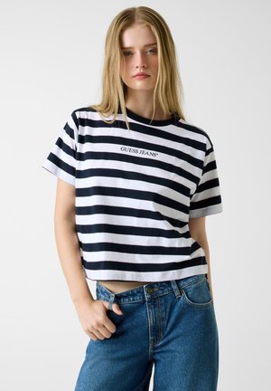 Zwart-wit gestreept cropped t-shirt met het "GUESS JEANS" logo, gemaakt van zachte katoen, gecombineerd met blauwe high-waisted spijkerbroek.