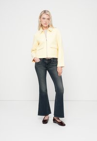 Κίτρινο κοντό μπουφάν με φερμουάρ μπροστά, δύο μπροστινές τσέπες, συνδυασμένο με σκούρο τζιν με καμπάνα και καφέ γυαλιστερά μπαλαρίνες. Απλό λευκό φόντο.