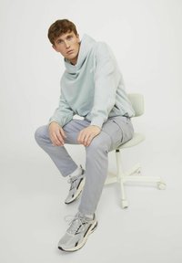 Hellblaue Kapuzenjacke, graue Cargo-Hose und graue Sneakers mit weißen Akzenten. Modell sitzt auf einem hellen Stuhl vor einem weißen Hintergrund.