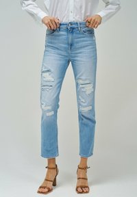 Vaqueros de mezclilla azul claro con pierna recta, detalles desgastados en las rodillas y una cintura de altura media. Combinados con tacones marrones de tiras.