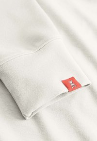 Lichtgrijze sweatshirtstof met een zachte textuur, voorzien van een rood label op de manchet met een wit grafisch ontwerp.
