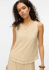 Beige mouwloos topje met een bloemenkantpatroon, voorzien van een ronde halslijn en een gebogen zoom. De stof lijkt licht en gestructureerd.