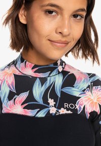Traje de neopreno con un patrón floral que presenta lirios rosas y hojas verdes sobre una base negra. Diseño de cuello alto con el logo "ROXY" en el pecho.