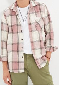 Heren flanellen overhemd in roze en wit ruitpatroon, met zwarte accenten, knopen aan de voorkant en een borstzak; gedragen over een wit T-shirt.