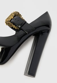 Zapato de tacón alto negro de charol con una prominente hebilla decorativa dorada y un tacón cuadrado y robusto. Textura suave y brillante con puntera redondeada.