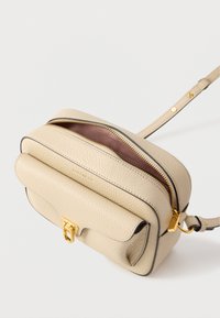 Béžová kožená kabelka přes rameno se zlatými kovovými doplňky, zavíráním na zip a texturovaným povrchem. Má zaoblený tvar a nastavitelný popruh.
