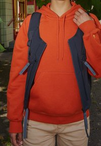 Persona con una sudadera naranja y pantalones beige, llevando un chaleco negro, sosteniendo una correa al aire libre en una zona sombreada cerca de la entrada de un edificio.