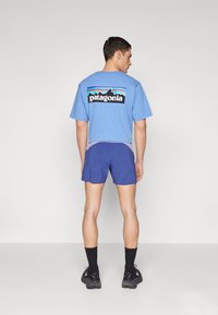 T-shirt de algodão azul com um logótipo gráfico nas costas, combinada com calções roxos e azuis. O modelo usa meias e sapatos pretos.