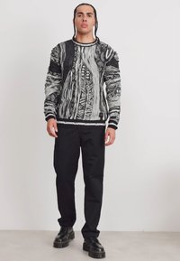 Schwarzer und weißer gemusterter Pullover mit strukturierten Details, Rundhalsausschnitt und gerippten Bündchen, kombiniert mit schwarzen Hosen und schwarzen Schuhen.