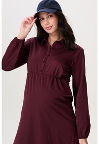 Frau in einem dunkelvioletten Langarmkleid und einer marineblauen Kappe, die mit ihrer rechten Hand den Schirm der Kappe gegen einen einfarbigen Hintergrund hält.