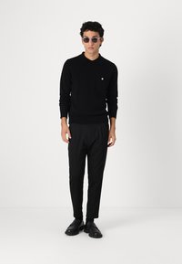 Maglione nero a maglia con colletto e piccola tasca, abbinato a pantaloni neri ben tagliati e scarpe nere. Gli accessori includono occhiali da sole grigi.