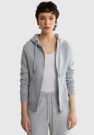 Tânăr cu păr scurt castaniu, purtând cercei aurii, un maiou alb și o hanorac cu fermoar gri deschis și pantaloni de trening asortați.