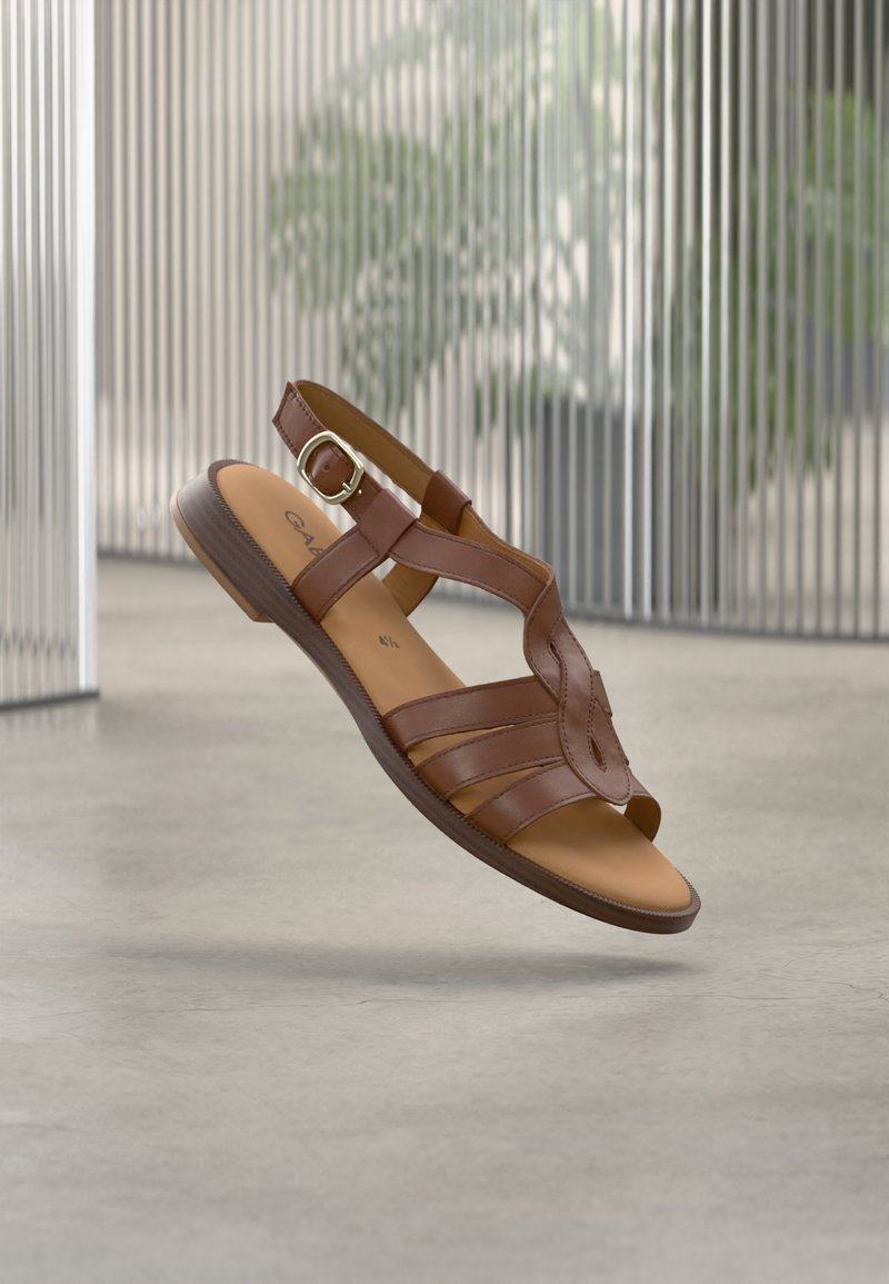 Sandale en cuir marron avec un design à lanières, bout ouvert et bride à la cheville, avec une boucle décorative et une semelle intérieure à texture lisse.