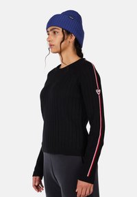 Schwarzer gerippter Pullover mit langen Ärmeln, der einen rot-weißen Streifenakzent entlang des Ärmels und ein Logo-Patch am Oberarm aufweist.