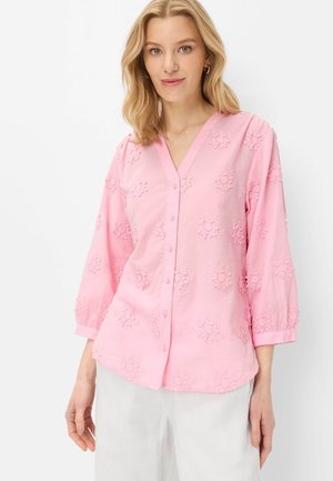 Femme portant un chemisier rose clair à boutons avec broderie florale et manches trois-quarts, associé à un pantalon blanc sur un fond uni.
