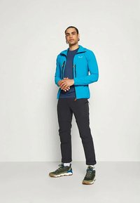 Blå lett jakke med glidelåslommer, sammen med en mørk skjorte og svarte bukser. Har grønne og grå tursko.