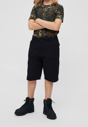 Kind draagt een groene camouflage-T-shirt, zwarte cargobroek en zwarte veterlaarzen, staat met gekruiste armen tegen een effen achtergrond.