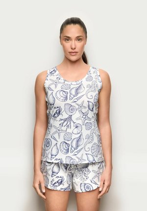 Frau trägt ärmelloses Oberteil und Shorts mit Muster aus blauen Meerestieren und Muscheln auf weißem Hintergrund, steht vor einer schlichten Wand.