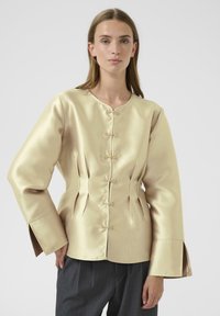 Satin blouse in light gold with a round neckline, diagonal pleats, and decorative knot closures. Features wide sleeves and a tailored fit.