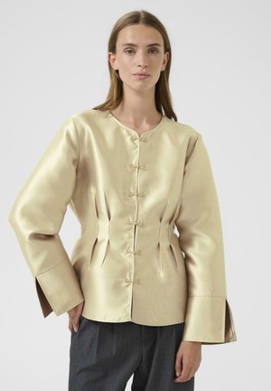 Blouse en satin doré clair avec un col rond, des plis diagonaux et des fermetures décoratives en nœud. Dispose de manches larges et d'une coupe ajustée.