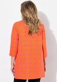 Långärmad tunika i starkt rosa med ett orange abstrakt mönster, mjukt tyg, rundad nederkant och sidoklipp, sedd bakifrån.