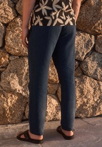 Dark blue, straight-leg pants with subtle pinstripes, made of lightweight fabric, paired with brown sandals and a patterned top against a rocky wall.