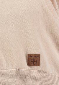 Maglione a maglia beige chiaro con un tessuto testurizzato. Presenta un'etichetta in pelle marrone con il nome del marchio "KEY LARGO" e il logo ricamato sul davanti.