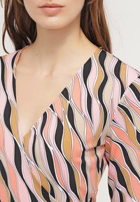 Vestido tipo wrap con un escote en V profundo, fabricado en un tejido suave con patrones ondulados en rosa, durazno, negro y beige. Primer plano de la parte superior de la prenda.