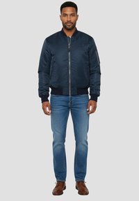 Blouson aviateur bleu marine avec fermeture éclair avant, poignets côtelés, poches latérales et texture lisse, associé à un jean en denim bleu et des chaussures marron.