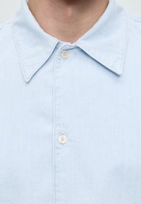 Gros plan d'un col de chemise bleu clair à boutons avec deux boutons blancs fermés sur le cou et le haut de la poitrine d'une personne.