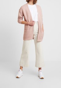 Helles rosa gestrickte Strickjacke mit offener Vorderseite, kurzärmelig und zwei Taschen auf der Vorderseite, kombiniert mit cremefarbenen weiten Hosen und weißen Turnschuhen.