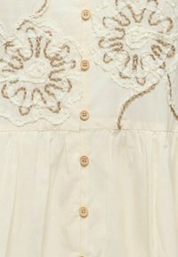 Tissu crème avec boutons beige et broderie florale en relief en fil blanc et marron clair sur le panneau avant du vêtement.
