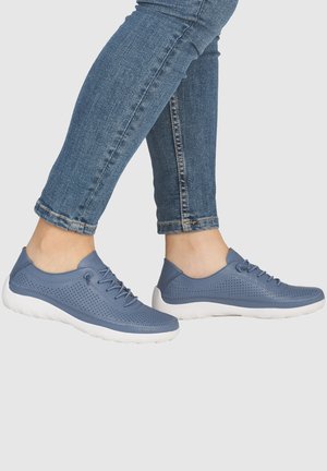 Person trägt blaue perforierte Ledersneaker mit weißen Sohlen und hochgekrempelte blaue Jeans vor hellem Hintergrund.