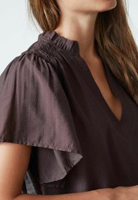 Blusa marrone scuro con scollatura a V, colletto arricciato e maniche a volant. Il tessuto ha un aspetto morbido e testurizzato.