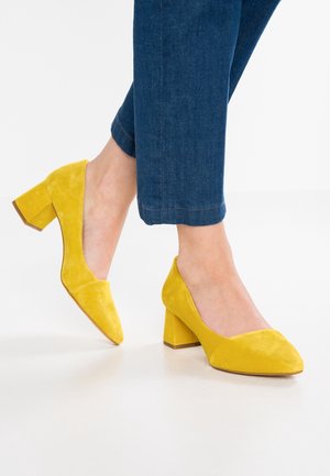 Żółte zamszowe buty na słupku z ostrym noskiem. Obcas jest gruby, co zapewnia stabilność oraz teksturowane wykończenie.