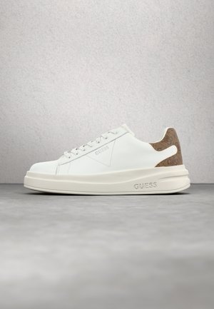 Białe niskie sneakersy Guess z kremową podeszwą, białymi sznurówkami i brązową teksturowaną naszywką na pięcie na szarej powierzchni i jasnoszarym tle.