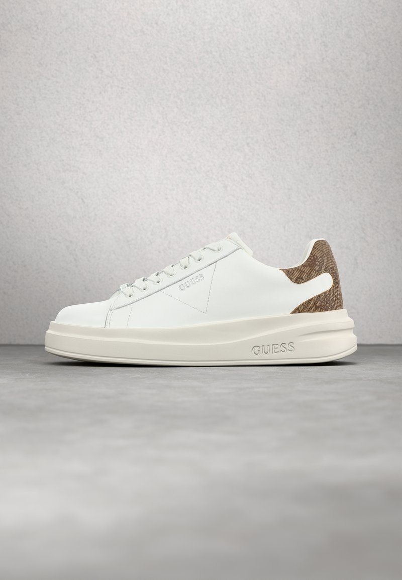 Baskets basses blanches Guess avec semelle crème, lacets blancs et patch de talon brun texturé sur surface grise et fond gris clair.