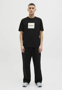 Sort bomulds t-shirt med et firkantet logo i beige og gul på brystet, parret med løse sorte bukser og grå træningssko.