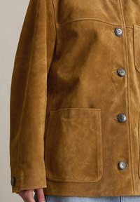Braune Wildlederjacke mit Knopfverschluss, die zwei große Fronttaschen und eine glatte Textur aufweist, von der Taille aufwärts gezeigt.