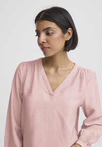 Růžová halenka s dlouhými rukávy a výstřihem do V, s nařasenými detaily na ramenech a manžetách. Hladká textura s mírně krčeným efektem.