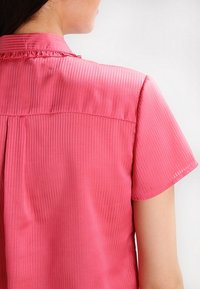 Rosa kurzärmliges Hemd mit vertikalen Streifen, einem Kragen mit Rüschendetails und einer glatten Stofftextur. Rückansicht dargestellt.