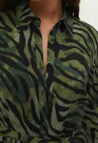 Camicia con bottoni in un motivo a strisce tigre verde, nera e oliva; presenta un colletto, bottoni verdi e un tessuto morbido e strutturato.