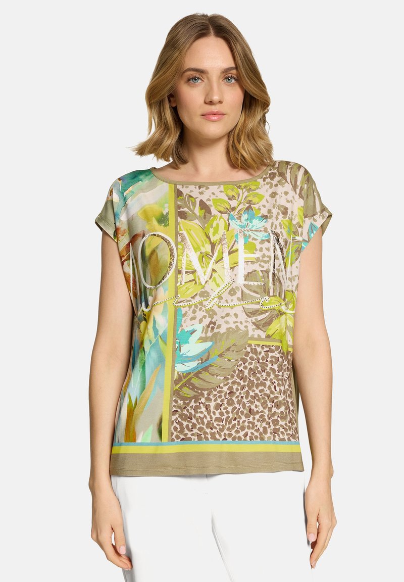 Femme portant un haut à manches courtes multicolore avec des motifs floraux et animaliers, et un pantalon blanc, sur un fond uni.