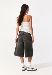 Culottes en denim noir avec ourlet effiloché, associés à un top blanc moulant sans bretelles et des baskets blanches. L'ensemble présente une coupe décontractée et ample.