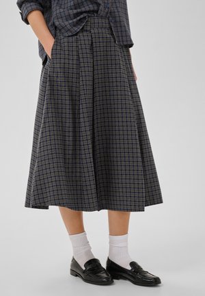 Personne portant une jupe midi à carreaux bleue, une chemise à carreaux assortie, des chaussettes blanches et des mocassins noirs, debout les mains dans les poches.