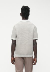 Muž s tmavou pokožkou a dredy, který má na sobě světle šedé krátké pletené tričko a světle hnědé kalhoty, stojí zády k bílému pozadí.