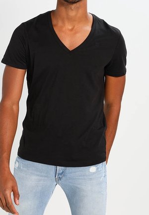 Mann trägt ein eng anliegendes schwarzes V-Ausschnitt-T-Shirt und hellblaue, abgenutzte Jeans, steht vor einem schlichten weißen Hintergrund.