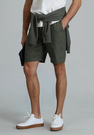 Shorts vert olive en tissu léger, avec une coupe décontractée et des poches latérales. Baskets blanches avec une semelle beige et un pull en tricot gris noué à la taille.