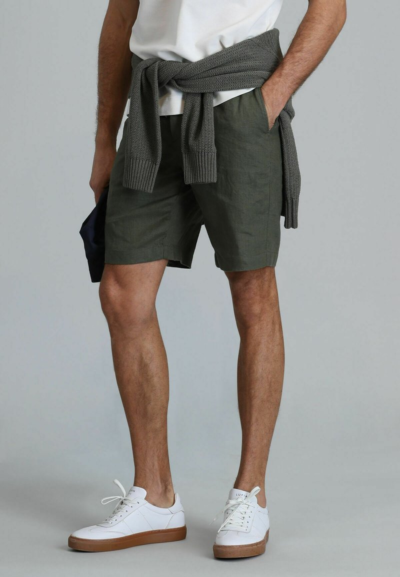 Olijfgroene shorts van lichtgewicht stof, met een casual pasvorm en zijzakken. Witte sneakers met een tan-zool en een grijze gebreide trui om de taille geknoopt.